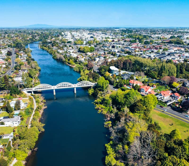 An aerial drone view looking down the wide blue Waikato River as it flows through the city of Hamilton, spanned by the distinctive three-arched white Fairfield Bridge. Credit: Josh Tere - Unsplash