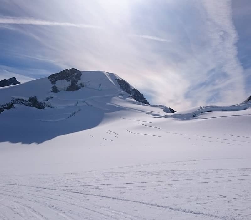 A vast, pristine snowfield on the upper reaches of Franz Josef Glacier, with deep crevasses visible on the mountain slopes under a bright sun and wispy clouds. Credit: Fine Tours Group