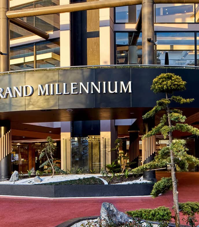 Modern entrance of Grand Millennium hotel with glass walls, columns, plants, and red carpet driveway. Credit: Grand Millennium Auckland