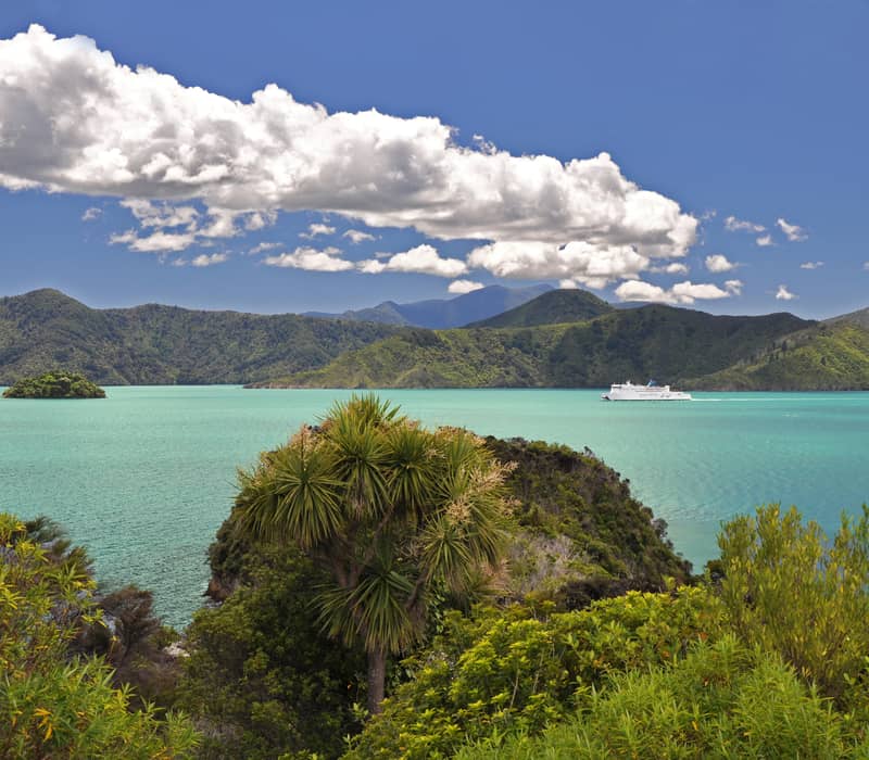 White ferry on turquoise bay water with green islands and hills, under blue sky with clouds. Credit: Fine Tours Group - Photographer unknown