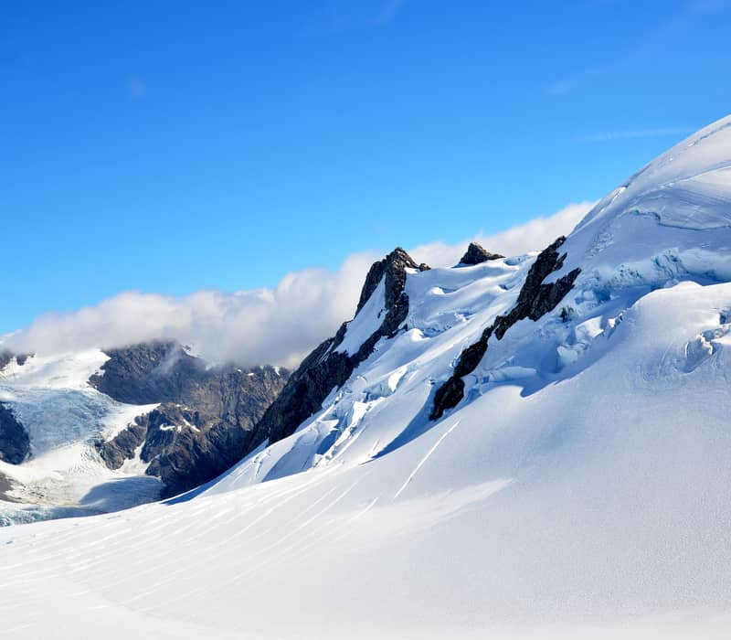 A bright, sunlit view of a steep, snow-covered mountain ridge with blue ice crevasses, set against a vibrant blue sky in the Southern Alps. Credit: Fine Tours Group