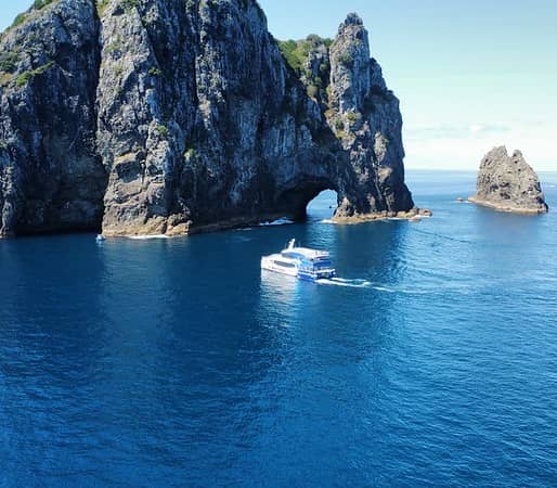 Boat sailing near rocky island with natural arch in blue ocean under clear sky. Credit: Explore Ltd