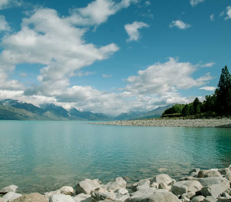 The bright turquoise waters of Lake Pukaki with a rocky stone shoreline in the foreground and snow-dusted mountains under a cloudy blue sky. Credit: Delphine Ducaruge - Unsplashed