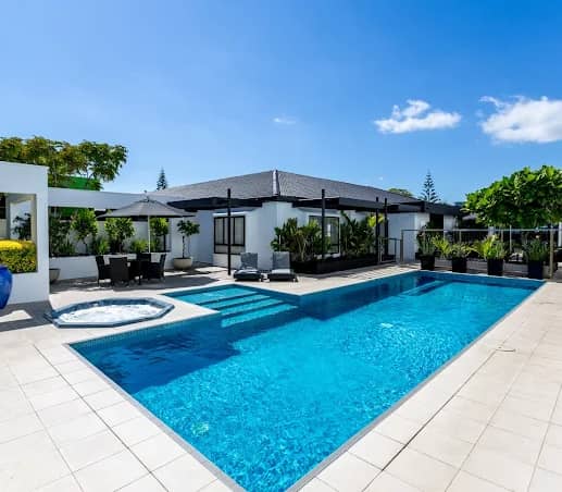 Modern outdoor swimming pool with patio, lounge chairs, umbrella, and plants under a bright blue sky. Credit: Leisure Time Travel