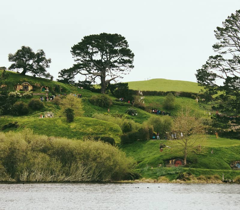 A wide landscape view across a lake looking at the rolling green hills of the Hobbiton Movie Set, dotted with several quaint Hobbit holes, large trees, and walking tourists. Credit: Raelle Cameron - Unsplash