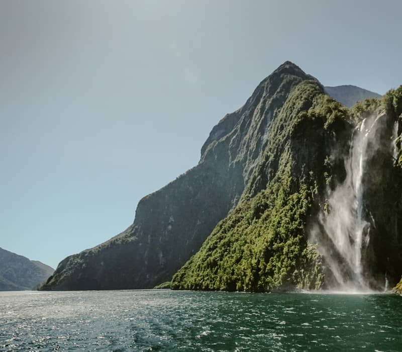 A massive waterfall cascading down a steep, lush green mountain cliff into the dark green waters of Milford Sound. Credit: Sandro Scalco - Unsplash