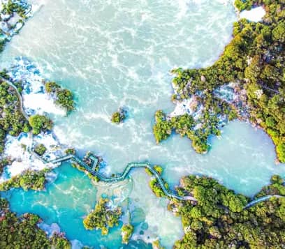 An aerial bird's-eye view of a winding wooden boardwalk crossing through vibrant turquoise and pale green geothermal pools surrounded by dense green native bush. Credit: Rotorua NZ - Media Library