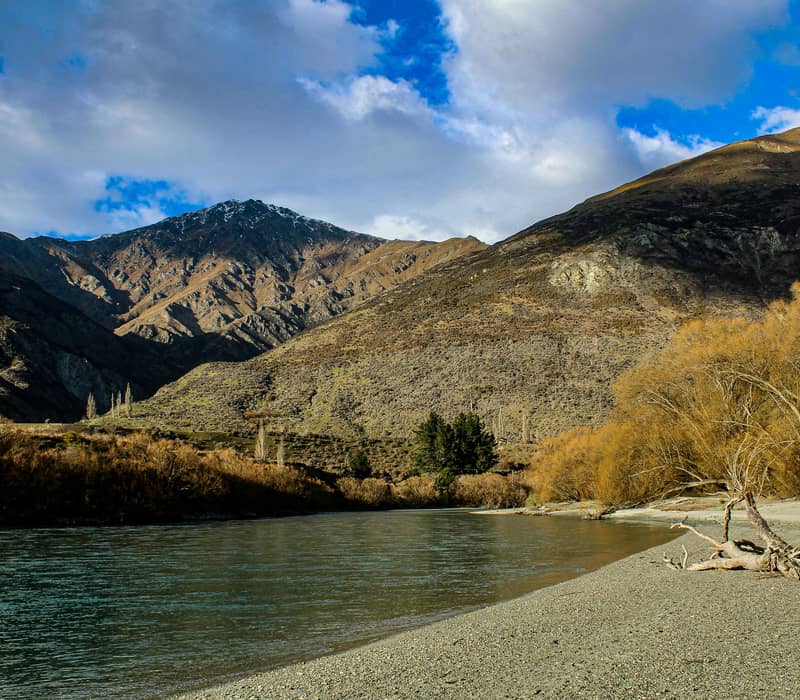 A scenic view looking down a wide, greenish river flowing past a grey pebble shoreline with golden autumn trees, beneath massive rugged mountain peaks under a cloudy blue sky. Credit: Mikyla Eden - Unsplash