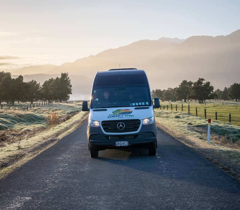 A travel van drives down a country road with mountains and trees in the background at sunrise. Credit: Cheeky Kiwi Travel