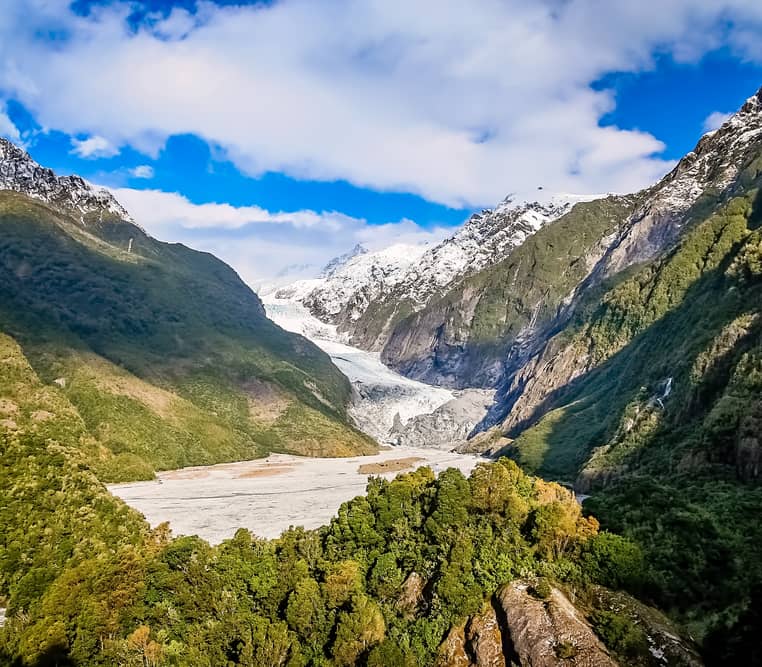A wide valley view showing the white ice of Franz Josef Glacier descending between steep, green forest-covered mountains under a blue sky with white clouds. Credit: Fine Tours Group - Photographer unknown