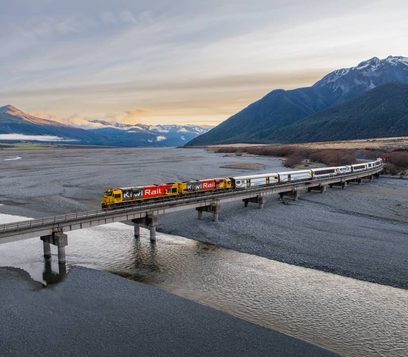Aerial view of a KiwiRail train on a bridge over river with mountains and clouds at sunset. Credit: Kiwi Rail