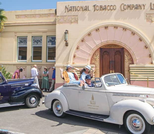 A group of people in 1930s vintage clothing waving from a white convertible and a dark blue vintage car parked in front of the ornate, peach-toned arched entrance of the National Tobacco Company building. Credit: Leisure Time Travel