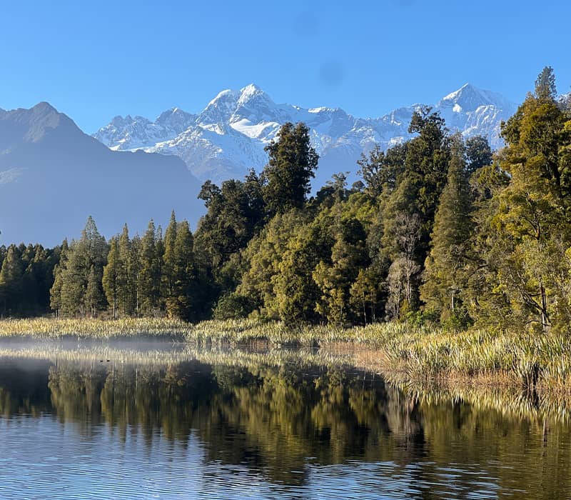 Calm lake with forest trees and snow-covered mountains reflected on water under clear sky. Credit: Cheeky Kiwi Travel