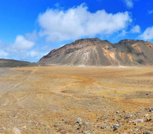 A wide panoramic view of the Tongariro Alpine Crossing showing hikers walking through a barren, rocky volcanic crater with the towering cone of Mount Ngauruhoe in the background under a blue sky. Credit: Fine Tours Group - Photographer unknown