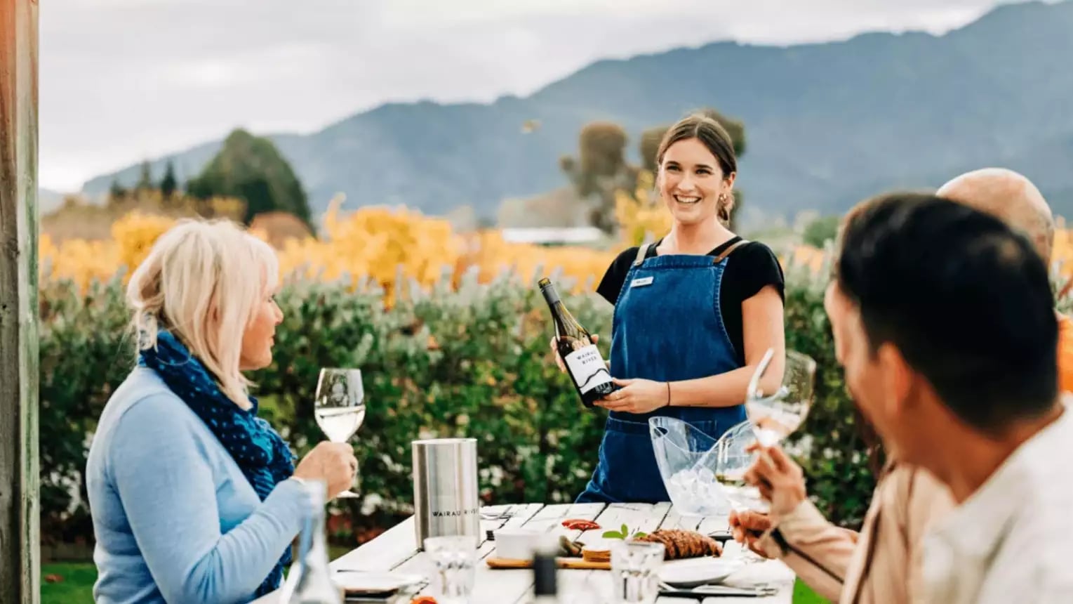 People enjoying outdoor wine tasting with a smiling host pouring wine at a vineyard with mountains behind. Credit: Great Journeys