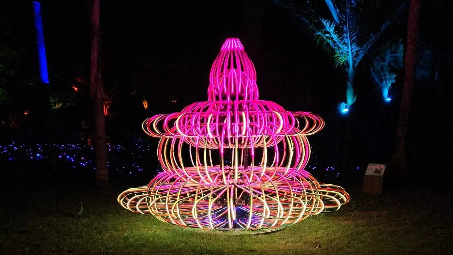 A vibrant neon light sculpture in the shape of a tiered fountain or pagoda, glowing with pink and orange lights in a dark park setting at night. Credit: Leisure Time Travel