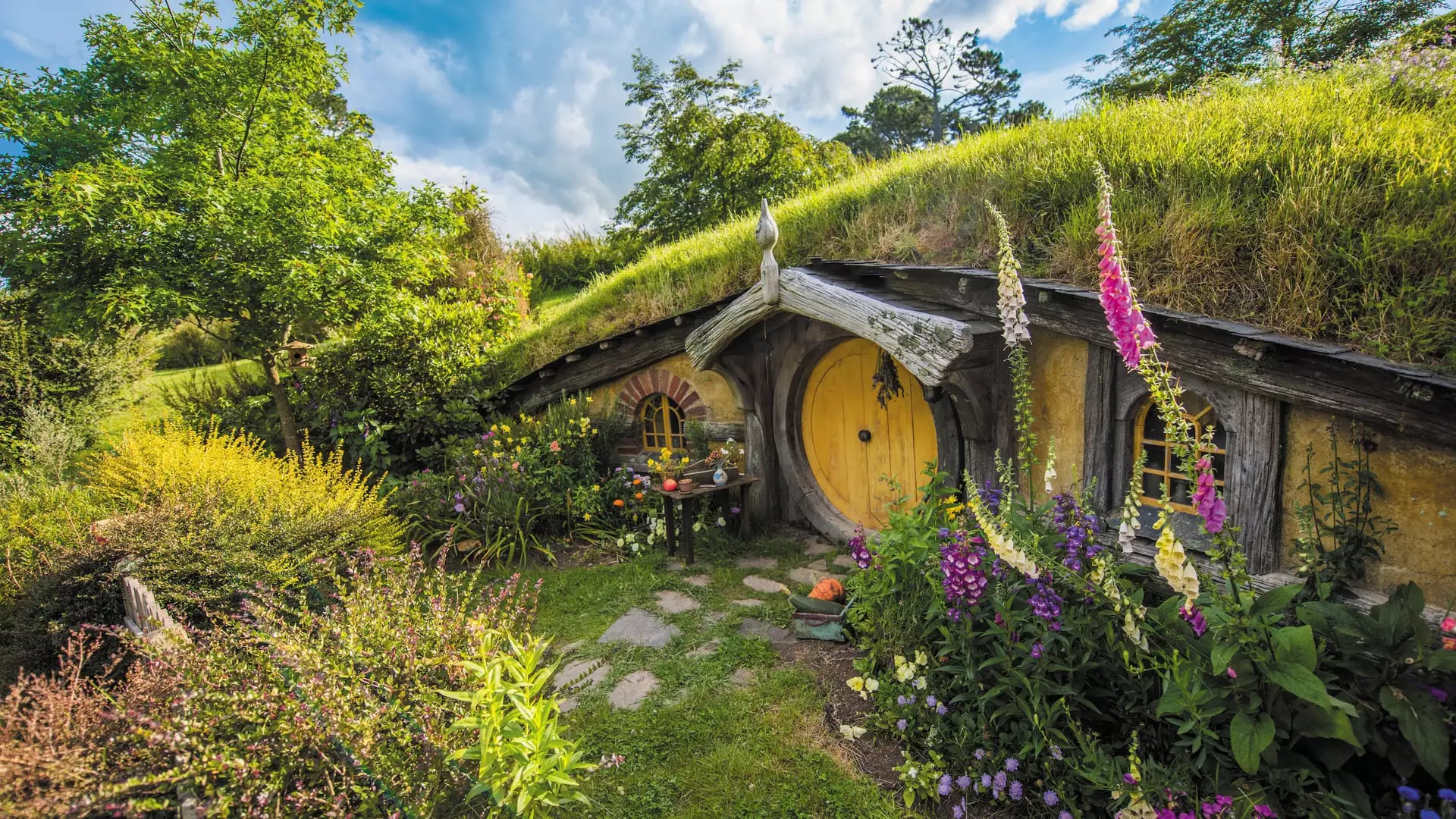 Hobbiton hobbit house entrance with round yellow door surrounded by colorful flowers and greenery under a blue sky. Credit: New Zealand Fine Touring