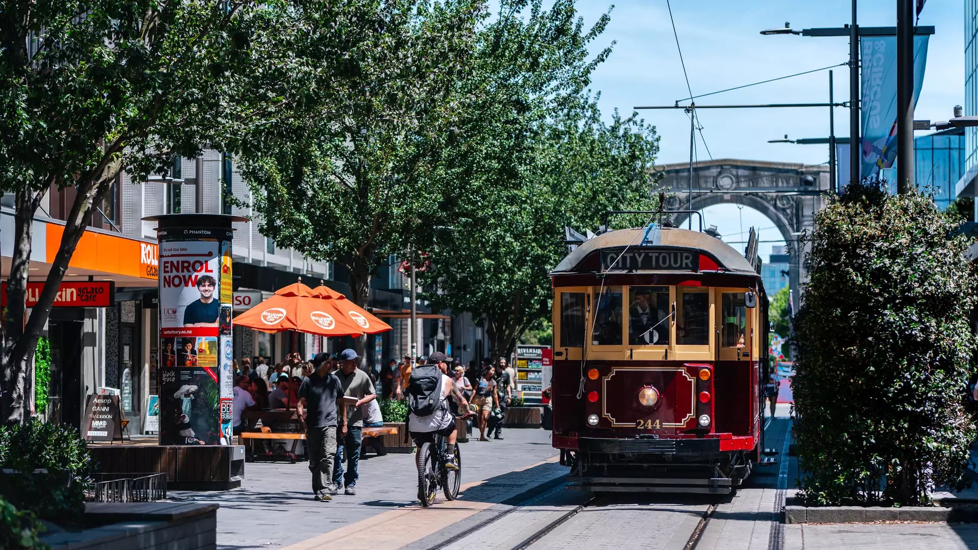 Vintage red and yellow tram on tracks with people walking and cycling in a tree-lined city mall on a bright day