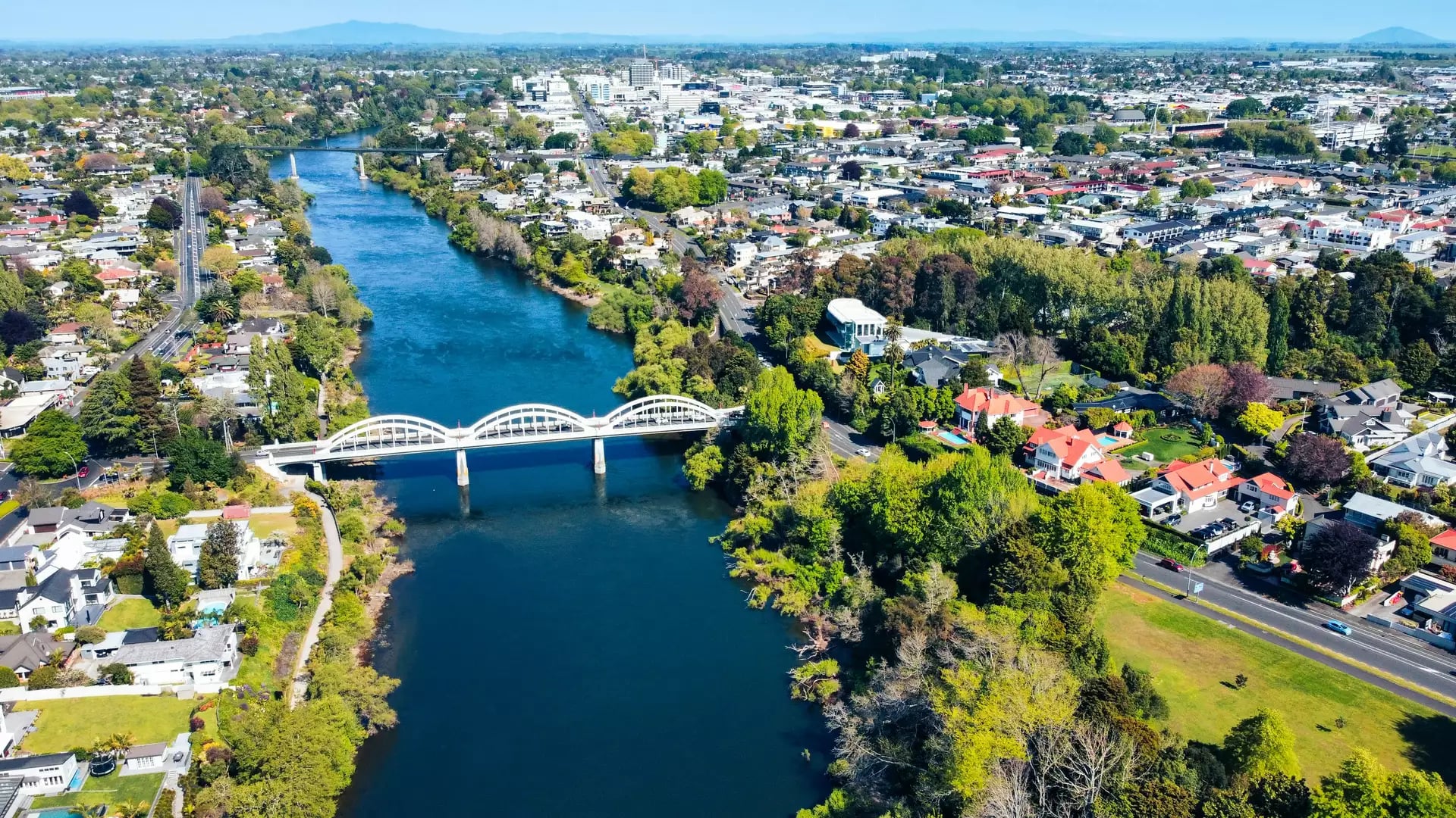 An aerial drone view looking down the wide blue Waikato River as it flows through the city of Hamilton, spanned by the distinctive three-arched white Fairfield Bridge. Credit: Josh Tere - Unsplash