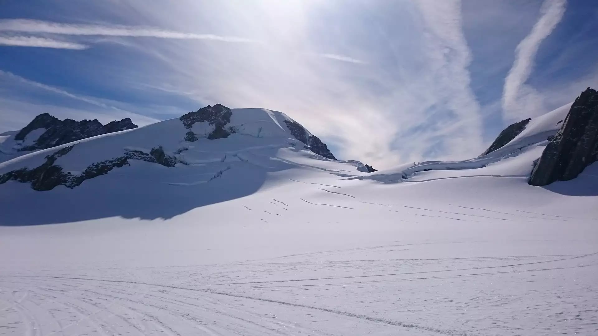 A vast, pristine snowfield on the upper reaches of Franz Josef Glacier, with deep crevasses visible on the mountain slopes under a bright sun and wispy clouds. Credit: Fine Tours Group