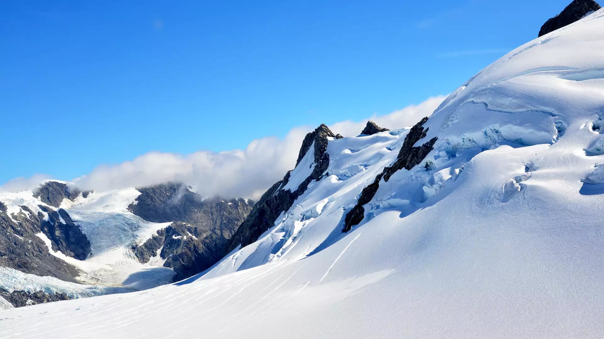 A bright, sunlit view of a steep, snow-covered mountain ridge with blue ice crevasses, set against a vibrant blue sky in the Southern Alps. Credit: Fine Tours Group
