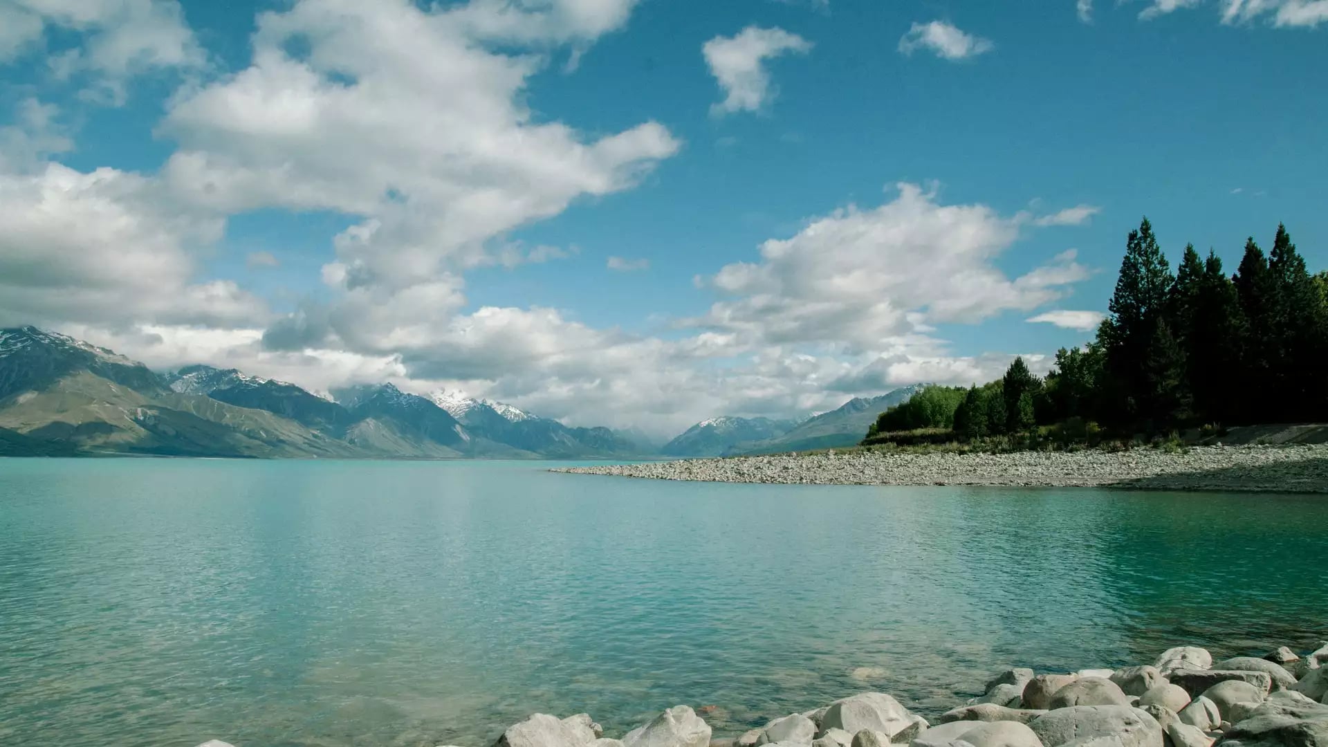 The bright turquoise waters of Lake Pukaki with a rocky stone shoreline in the foreground and snow-dusted mountains under a cloudy blue sky. Credit: Delphine Ducaruge - Unsplashed
