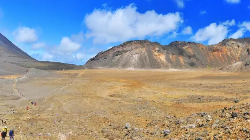 A wide panoramic view of the Tongariro Alpine Crossing showing hikers walking through a barren, rocky volcanic crater with the towering cone of Mount Ngauruhoe in the background under a blue sky. Credit: Fine Tours Group - Photographer unknown