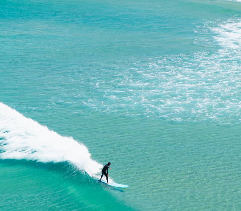 Surfer glides on a blue wave near a sandy beach on a sunny day.