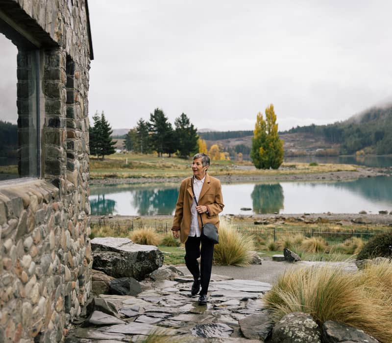 A woman in a tan coat walking along a stone path next to a historic stone church with a misty lake and mountains in the background. Credit: AAT Kings