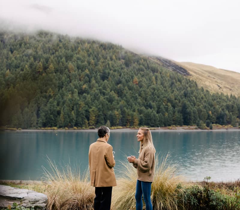 Two women with mugs standing in front of a bright blue lake and a steep, evergreen-covered mountain under low-hanging clouds. Credit: AAT Kings