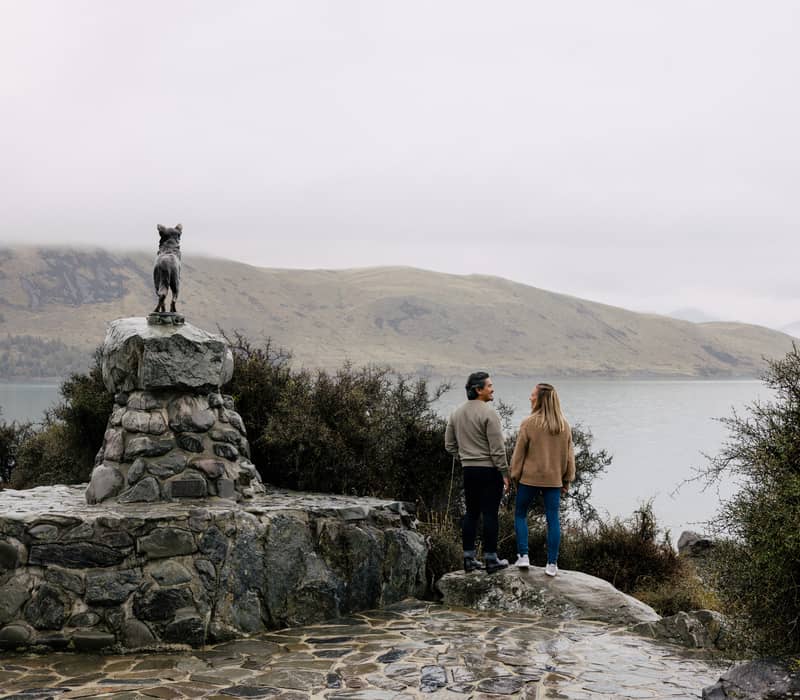 A couple standing on rocks near a bronze statue of a dog on a stone pedestal overlooking a wide, hazy lake and rolling hills. Credit: AAT Kings