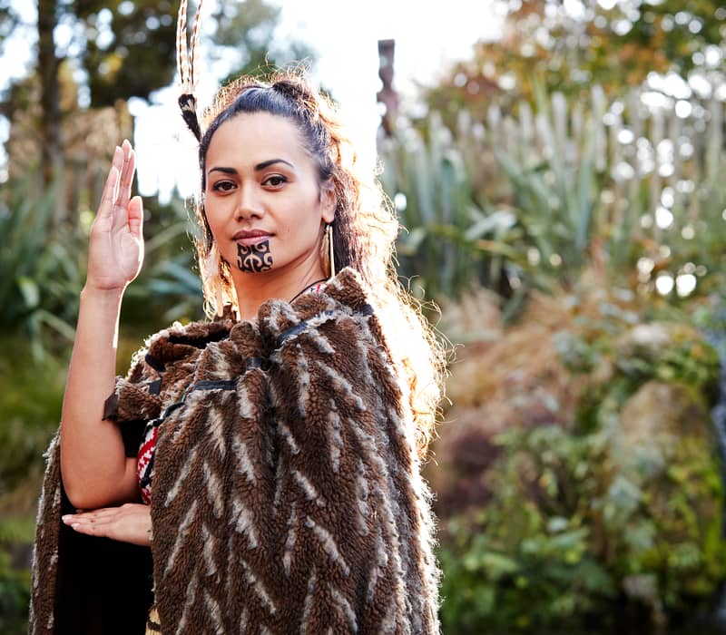 Maori woman with facial tattoo and traditional cloak standing near a waterfall in a forest. Credit: AAT Kings