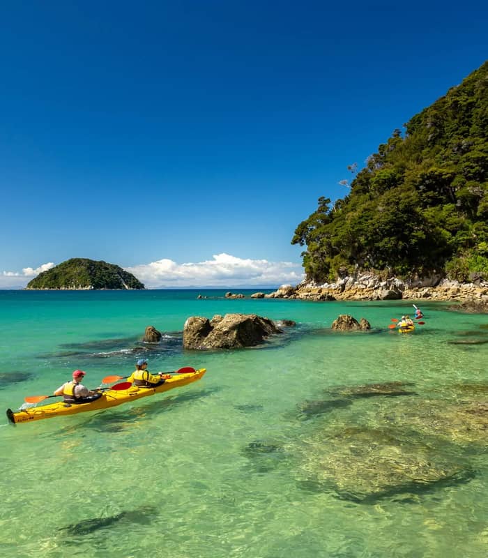Two people kayaking in clear turquoise waters near a rocky green coast under a blue sky. Credit: Abel Tasman Kayaks