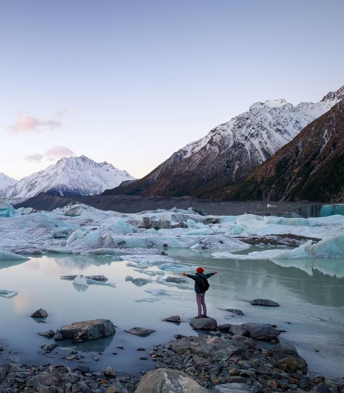 A person stands on a rocky shore with outstretched arms, looking over a glacial lake filled with icebergs under towering snow-capped peaks. Credit: Altitude Tours