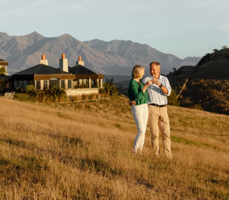 Couple standing on dry grass in front of a lodge with mountains at sunset.