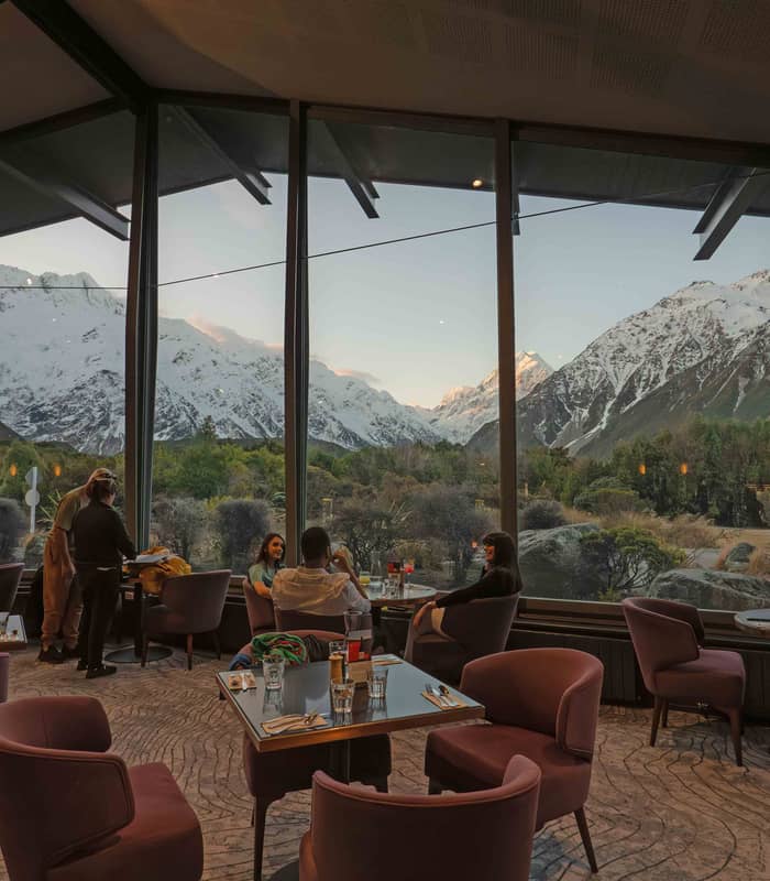 People dining inside a restaurant with large windows overlooking snow-covered mountain peaks.
