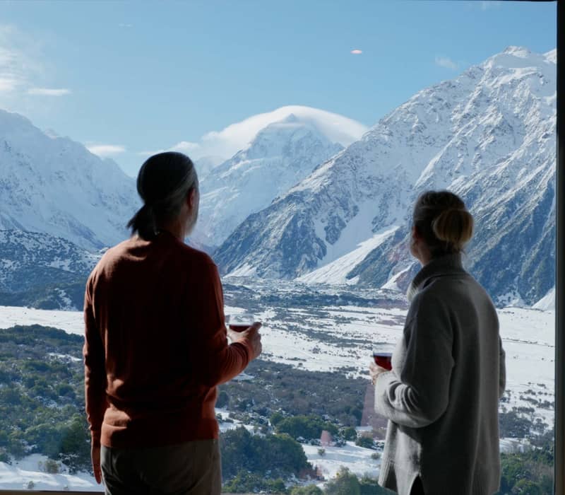 Couple drinking wine indoors looking at snowy mountain view through large window.