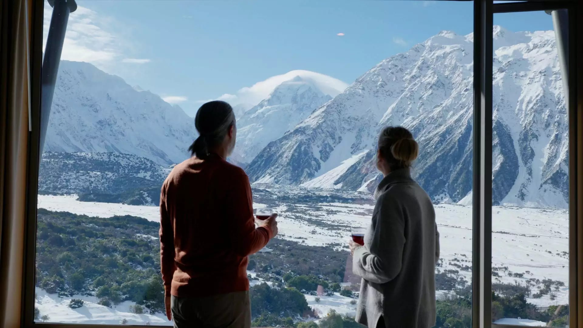 Couple drinking wine indoors looking at snowy mountain view through large window.