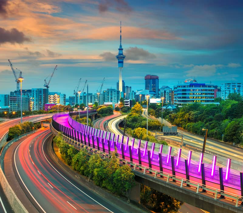 Auckland city skyline at sunset with Skytower and light trails on curved highways. Credit: G Adventures