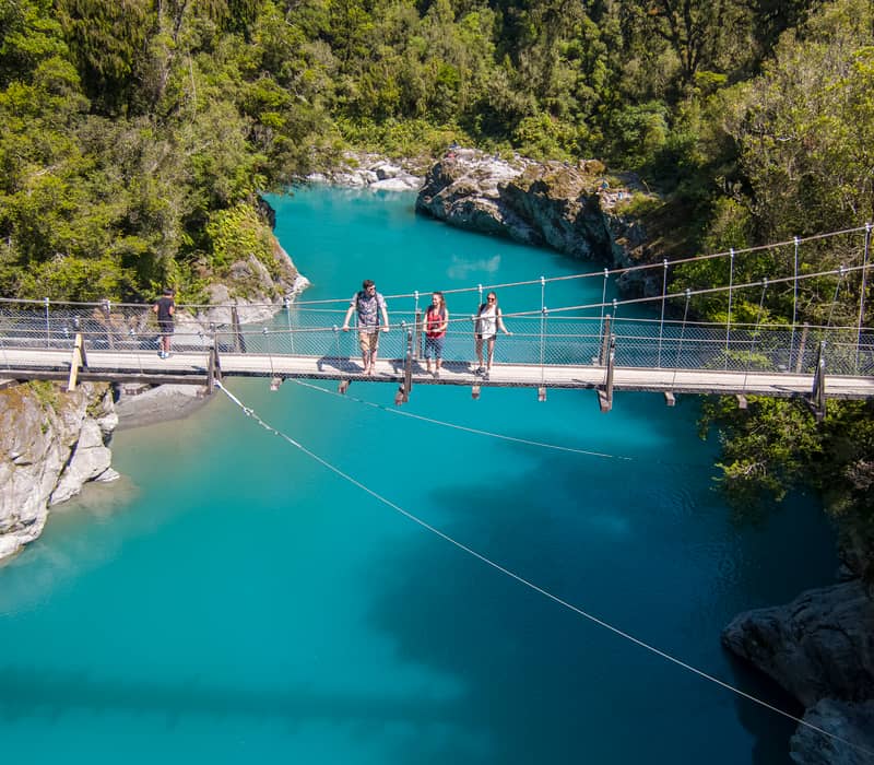 People crossing a suspension bridge over a bright blue river in a forested area. Credit: G Adventures