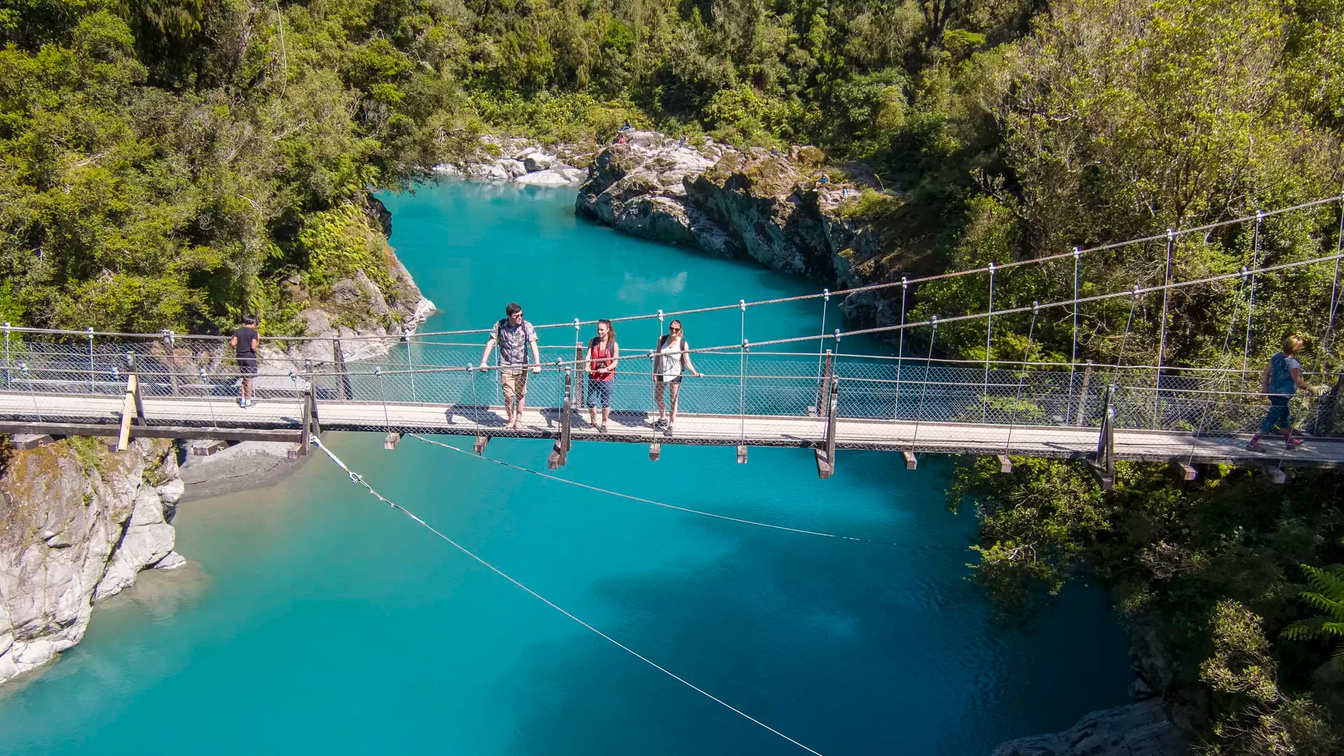People crossing a suspension bridge over a bright blue river in a forested area. Credit: G Adventures