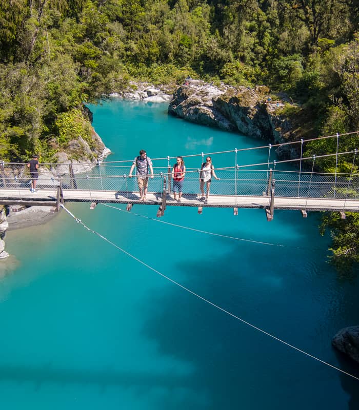 People crossing a suspension bridge over a bright blue river in a forested area. Credit: G Adventures