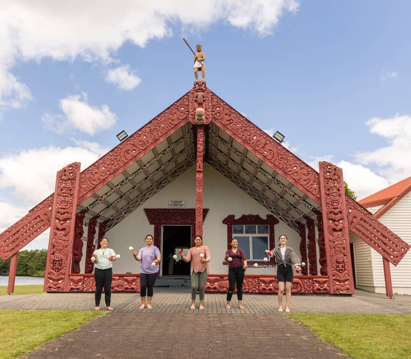 People barefoot in front of a red-carved Maori cultural house by a lake on a sunny day. Credit: G Adventures