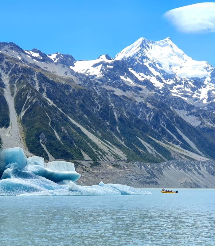 Boat with people near an iceberg on a lake with snow-covered Mount Cook in the background under a clear blue sky.
