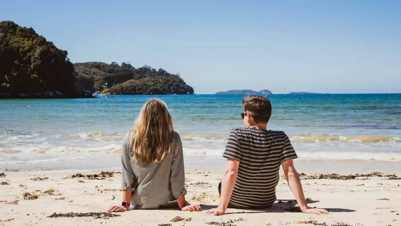 Two people sitting on a sandy beach looking out at calm ocean waters and forested islands. Credit: Glenorchy Air