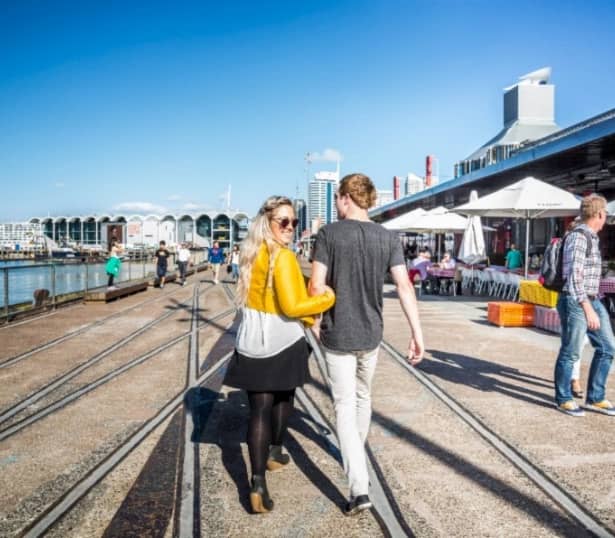 Couple walking on waterfront promenade with outdoor dining and buildings under clear blue sky. Credit: Great Journeys