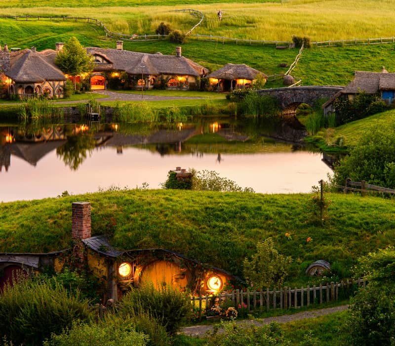 Hobbit houses with glowing windows and thatched roofs by a pond and stone bridge in green countryside at sunset. Credit: Cheeky Kiwi Travel