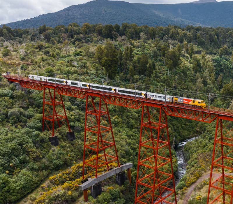 Train on red steel bridge over river amidst forest and mountains under cloudy sky Credit: Kiwi Rail