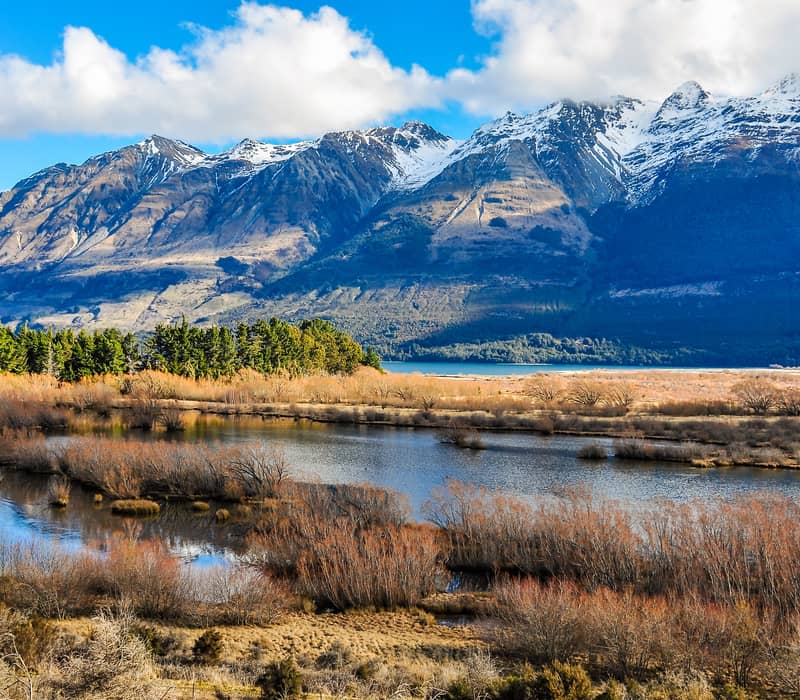 Snow-capped mountains and forest reflected in a calm lake under a partly cloudy sky.