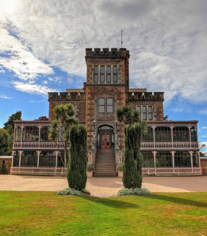 Front view of Larnach Castle with stone facade, two tall trees, and blue sky with clouds.